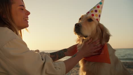 disparando de cerca: una chica rubia le desea a su perro un feliz cumpleaños en una playa soleada y le pone una gorra festiva