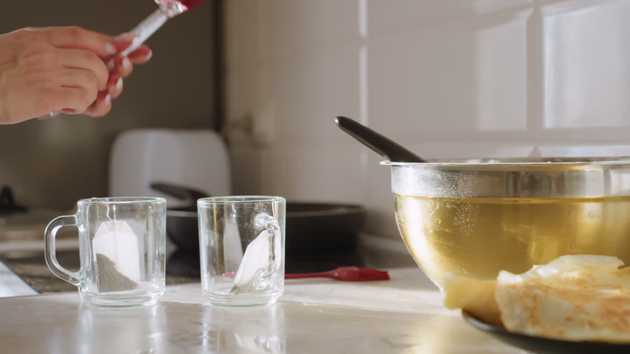 Крупный план руки, опускающей красный шпатель на кухонную столешницу рядом с двумя стеклянными чашками с чайными пакетиками внутри и большой металлической миской, наполненной жидкостью