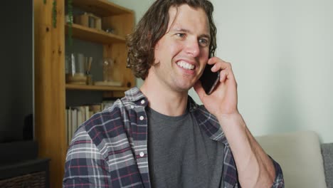 hombre caucásico feliz sentado en el sofá en la sala de estar hablando en el teléfono inteligente