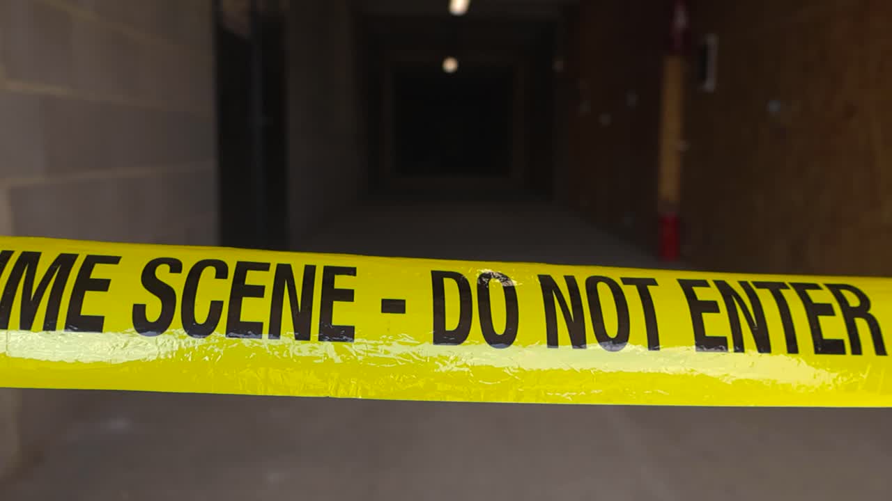 Close up view of a police line investigation forensics yellow colored Crime Scene tape pulled in front of a dark industrial hallway or corridor during day time. Concrete flooring and darkness visible