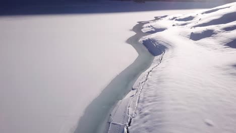 Close-aerial-view-over-the-shore-of-a-frozen-mountain-lake-to-a-huge-panorama-in-a-valley-in-KlÃ¶ntal,-Switzerland