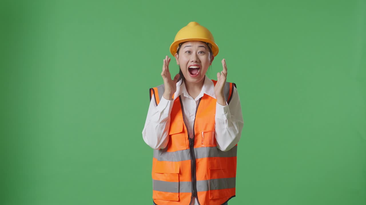 안전 헬을 입은 아시아 여성 엔지니어가 녹색 스크린 배경 스튜디오에 서서 카메라에 미소 짓고 와우라고 말합니다.