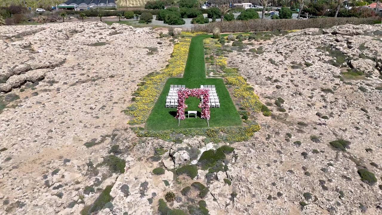 Un giardino è preparato per una cerimonia nuziale. Sedie bianche sono disposte in file e fiori sono posizionati davanti. Erba fresca e piante fiorite circondano l'area. Questo è un evento all'aperto e soleggiato.
