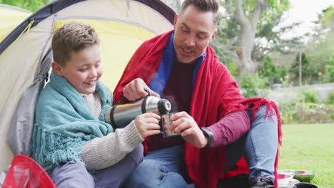 Happy-caucasian-father-with-son-sitting-in-tent-and-drinking-tea-in-garden