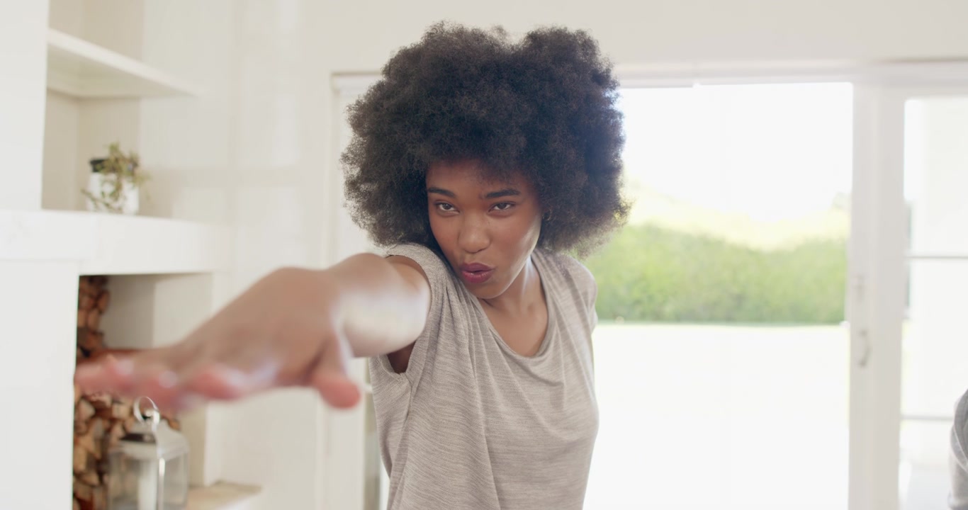 Mujer concentrada en casa, extendiendo el brazo hacia adelante, posando de forma juguetona con una camiseta gris junto a una puerta corrediza de cristal.