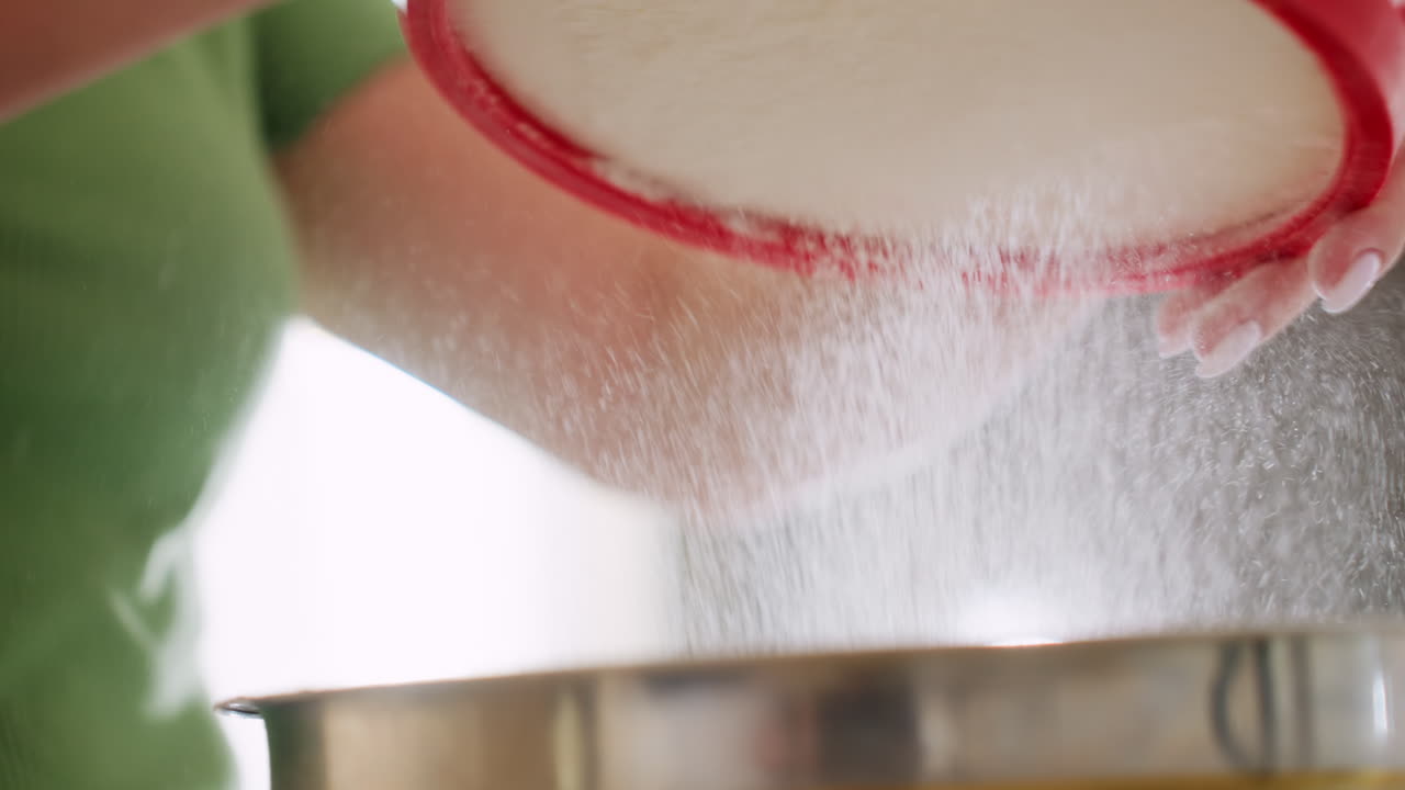 Close up of flour dust gently sifting into stainless bowl during baking preparation, fine particles floating in soft light as cook carefully filters ingredients for homemade recipe in kitchen