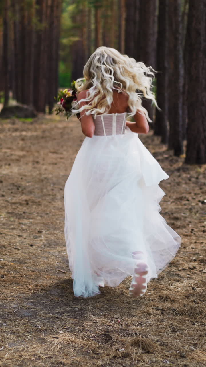Graceful young bride with long curly hair in elegant white wedding dress with fresh bouquet runs along pine forest ground path backside view slow motion