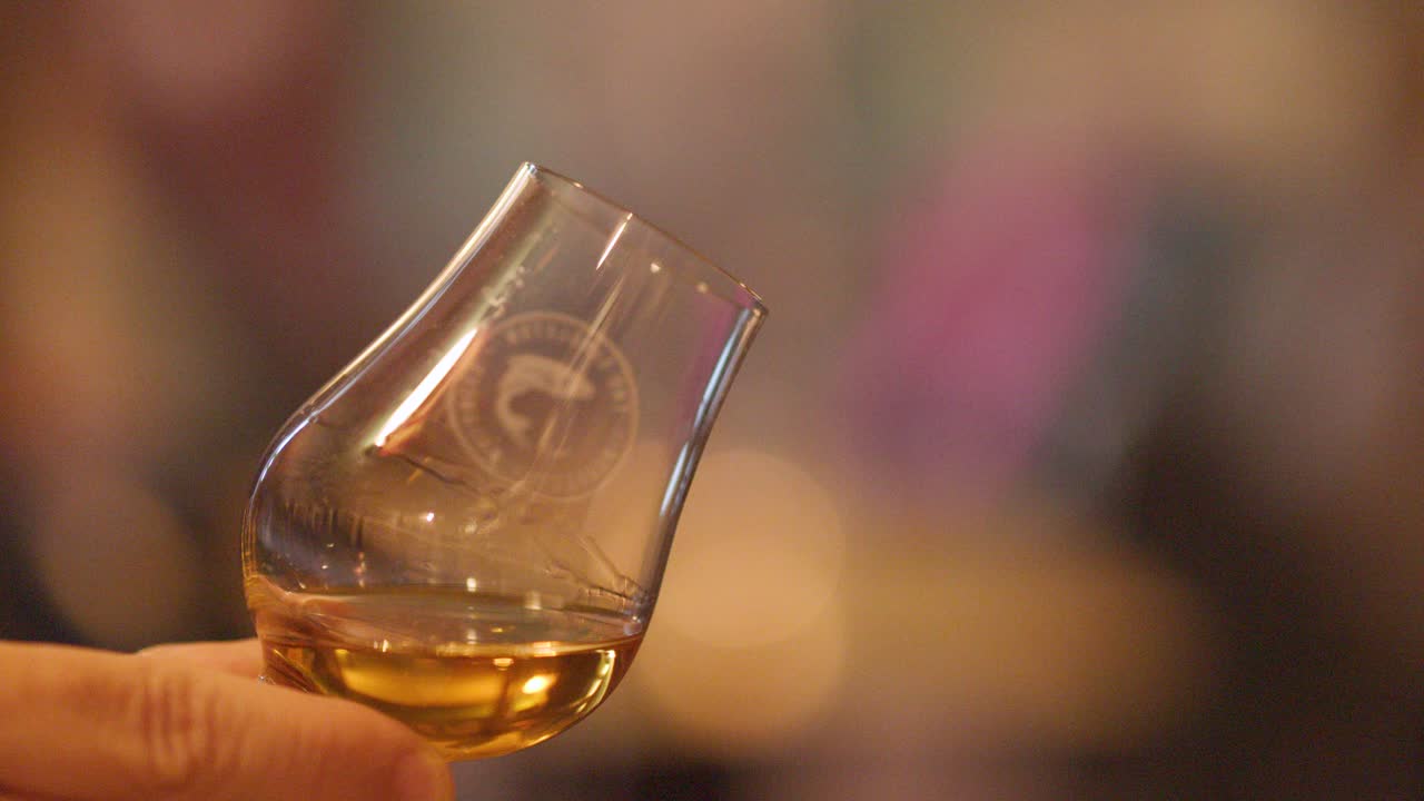 A hand gently swirls a whisky glass, highlighting amber liquid and reflections in warm, softly lit pub surroundings with a blurred, inviting background