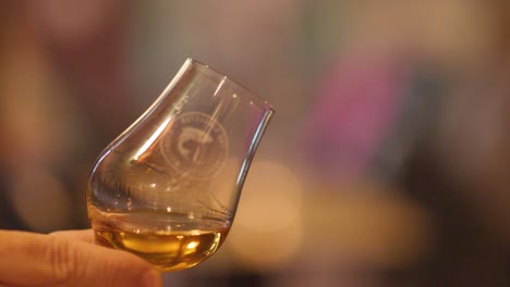a hand gently swirls a whisky glass, highlighting amber liquid and reflections in warm, softly lit pub surroundings with a blurred, inviting background
