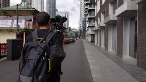 Young-caucasian-man-is-walking-with-his-backpack-and-camera-tripod-on-his-back-in-the-streets-of-the-business-district-Canary-Wharf-in-London-in-autumn
