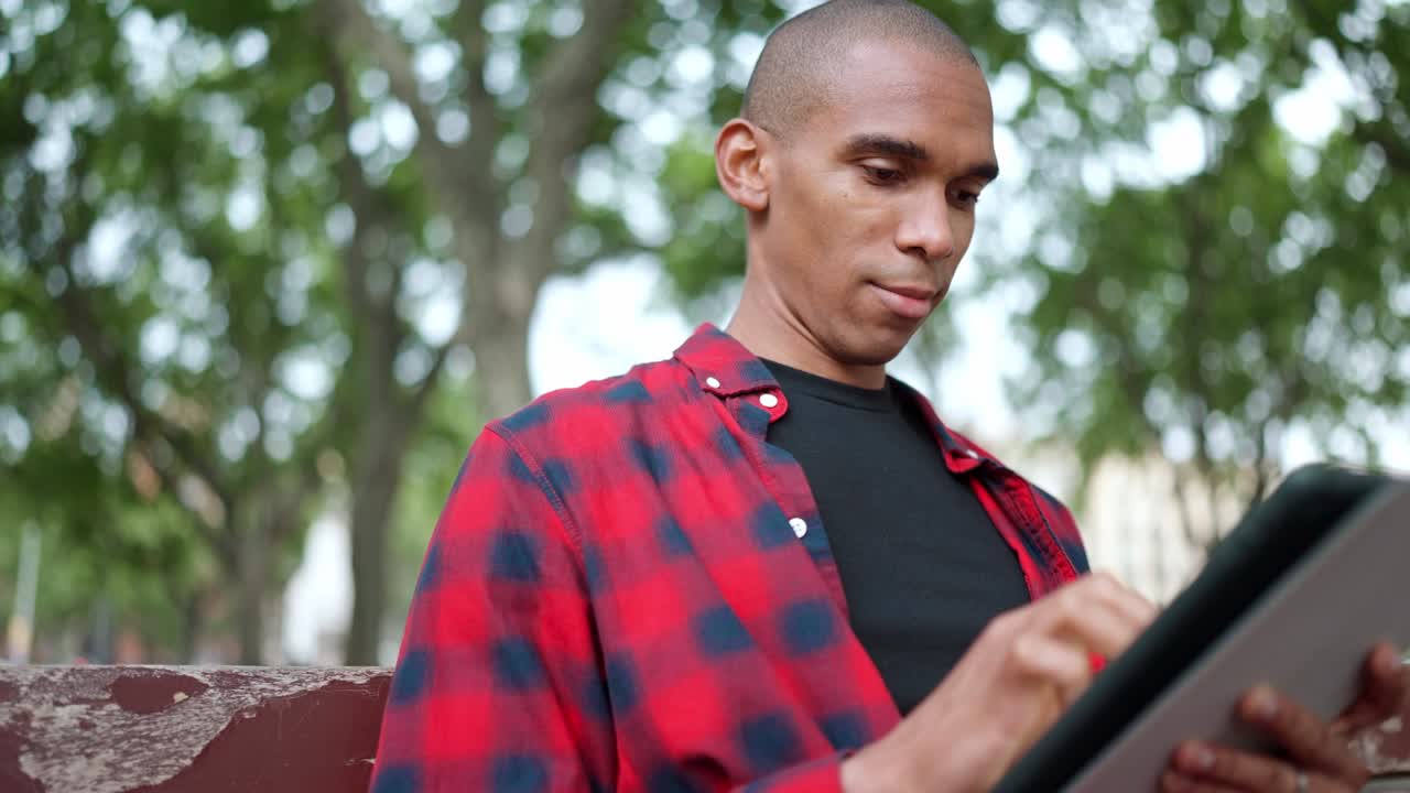 Pleased bald African man typing on digital tablet on bench