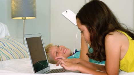 siblings sitting back to back and using digital tablet, laptop in bedroom