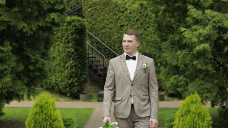 groom goes down the alley between bushes with a wedding bouquet to his beloved bride