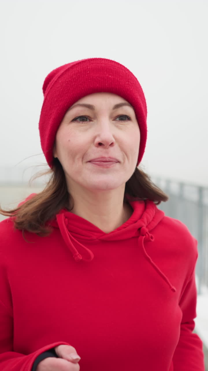 close up of woman jogging in red beanie and jacket along snowy pathway near iron railing in foggy winter park with benches and light pole distant bridge and snow covered trees and visible breath