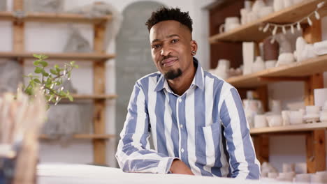 happy african american man with beard sitting at desk and smiling in pottery studio, slow motion