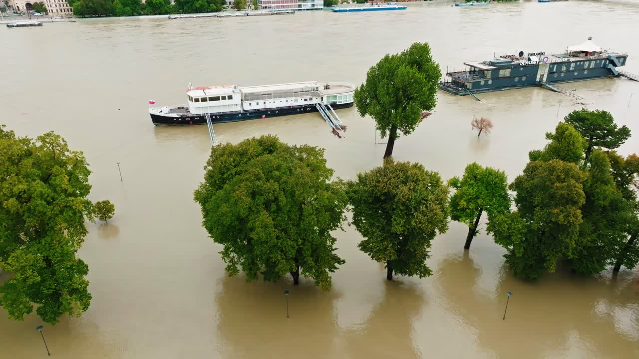 Aerial footage of extent of flooding along the Danube River, with water levels rising around trees along the riverbank. City buildings and boats are visible in the background. (4K)