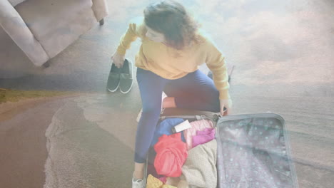 animation of happy caucasian woman sitting on floor packing suitcase over beach landscape