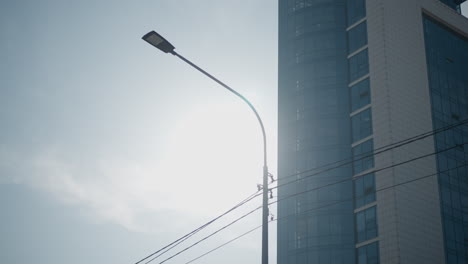 street light pole against blue sky with sun shining through, bright lens flare over modern glass office tower, utility wires crossing frame, clean minimal urban mood under crisp winter daylight