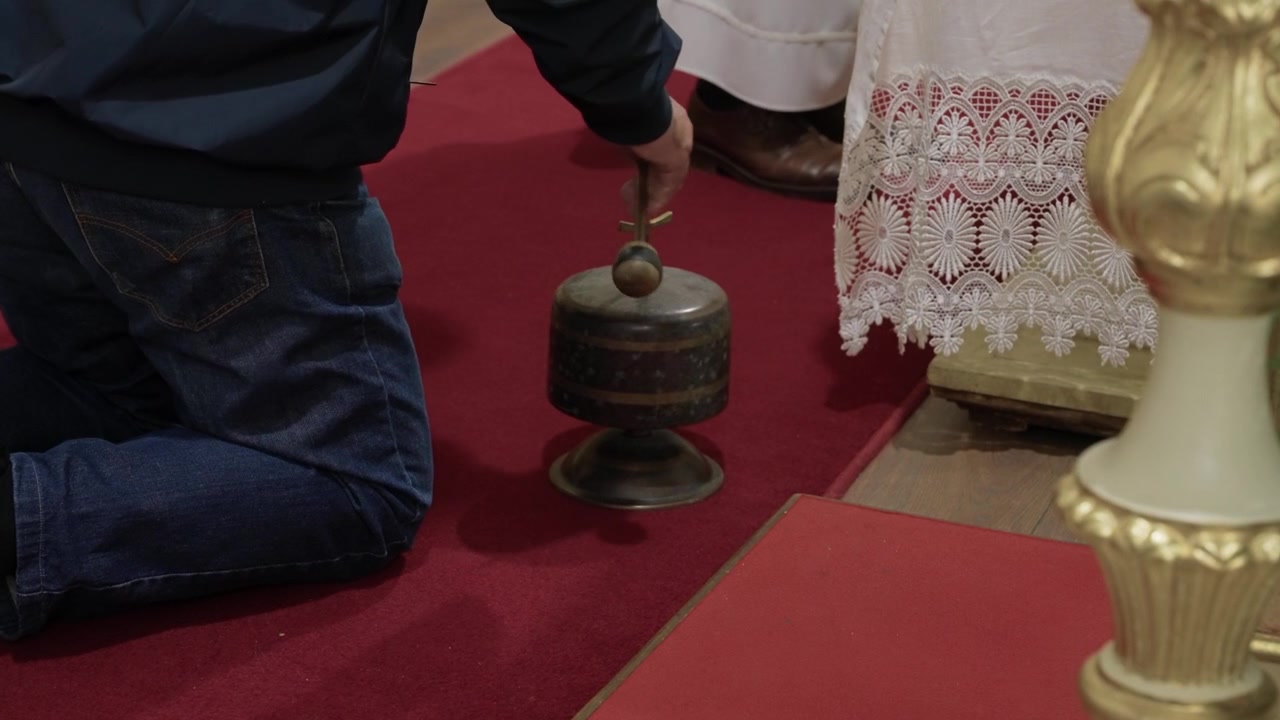 Hånd slår på ceremoniel metalklokke på rødt tæppe under kirkeceremoni.