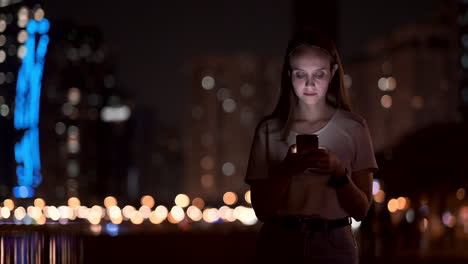 A-young-woman-looks-into-the-smartphone-and-writes-text-messages-on-the-Internet-against-the-backdrop-of-the-night-city.-Girl-businessman-working-on-vacation-remote-work-via-mobile-phone.-Gadget-addiction