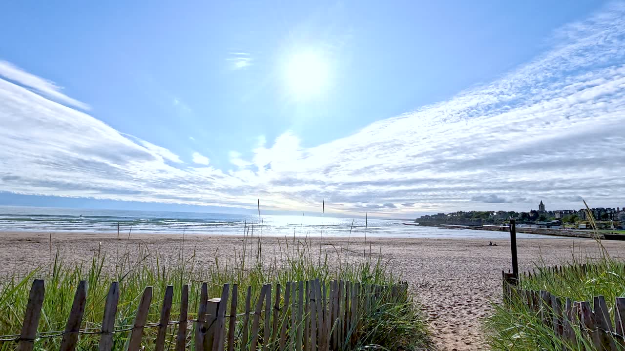Wide shot of sandy beach, wooden fence, and grassy dunes beneath bright blue sky