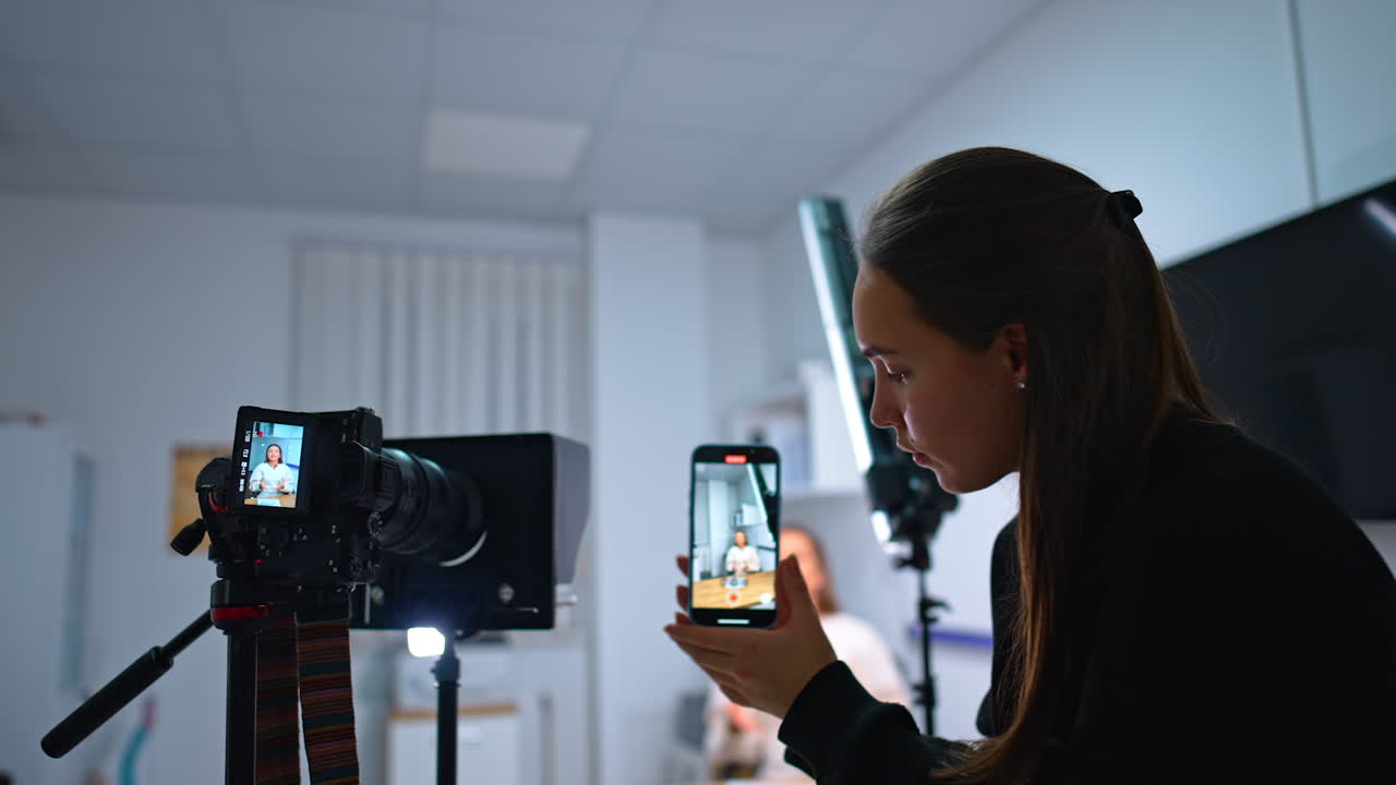 Recording female blogger on a professional camera and smartphone. Long-haired brunette holds her phone taking video of an influencer. Blurred backdrop.