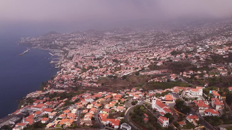 Imagens aéreas sobrevoando os telhados de terracota do Funchal e a extensa paisagem urbana, revelando vistas costeiras, colinas exuberantes e uma atmosfera nebulosa em um dia de luz difusa.