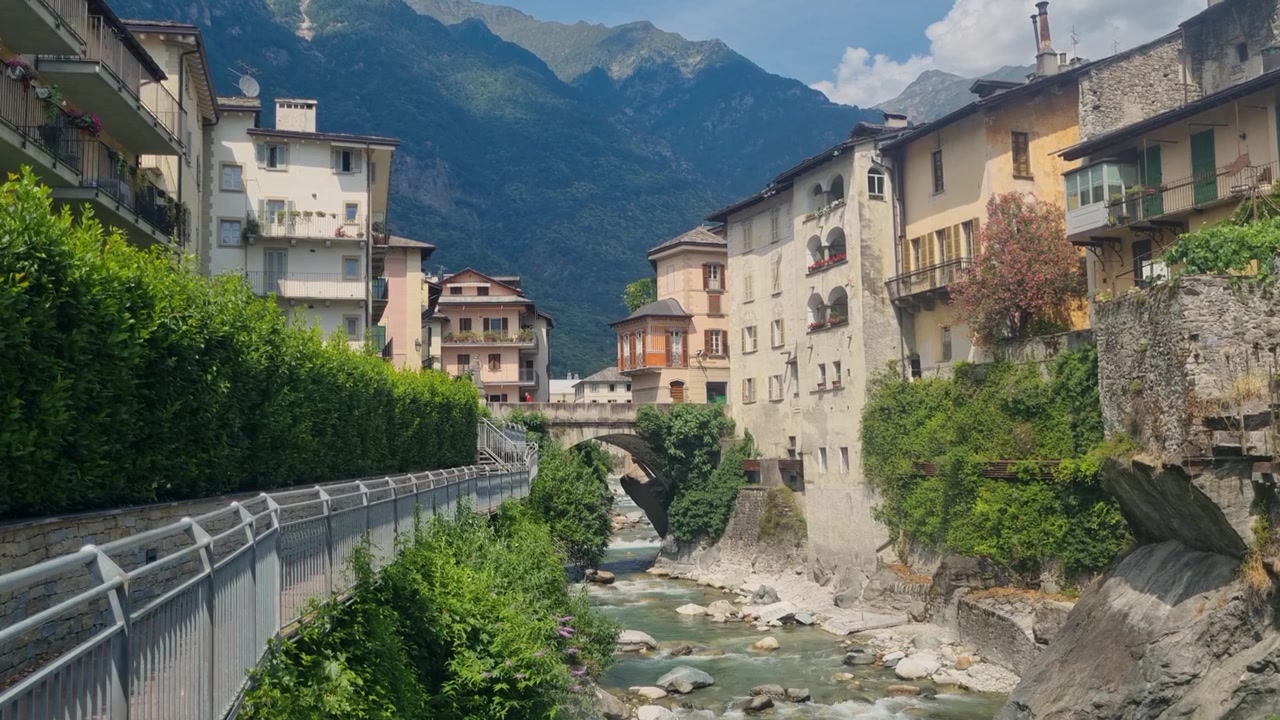 Splendida vista di Chiavenna in Lombardia, Italia, con la sua affascinante architettura lungo il fiume, il fiume Mera che scorre, la vegetazione mediterranea e le aspre Alpi Retiche che circondano questa storica città alpina