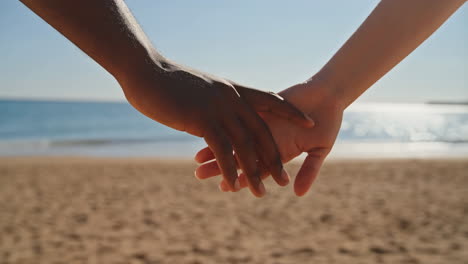 close-up of two people holding hands on a sandy beach with the ocean in the background