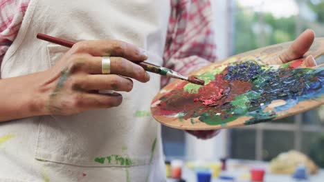 Hands-of-biracial-female-artist-in-apron-mixing-paints-on-palette-using-brush-in-studio,-slow-motion