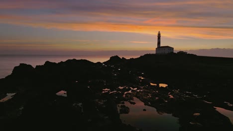 Paisaje-Panorámico-De-La-Puesta-De-Sol-Aérea-En-La-Costa-De-Menorca,-Horizonte-Del-Faro,-Cielo-Dorado-Y-Colorido-Con-Tonos-Rosados,-Isla,-Entorno-Rocoso-A-Orillas-Del-Mar