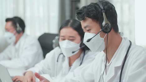 Close Up Of An Asian Man And A Woman Doctors Wearing Headsets And Masks Working As Call Center Agent Discussing Work While Their Colleague Is Speaking With Customer On The Call At The Office