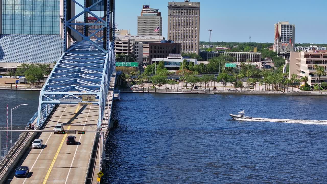 Boat on river below Bridge in Jacksonville City at sunny day. American flag on top of tower beside mirrored Wells Fargo Center Skyscraper against blue sky. Aerial zoom wide shot. Florida, USA.