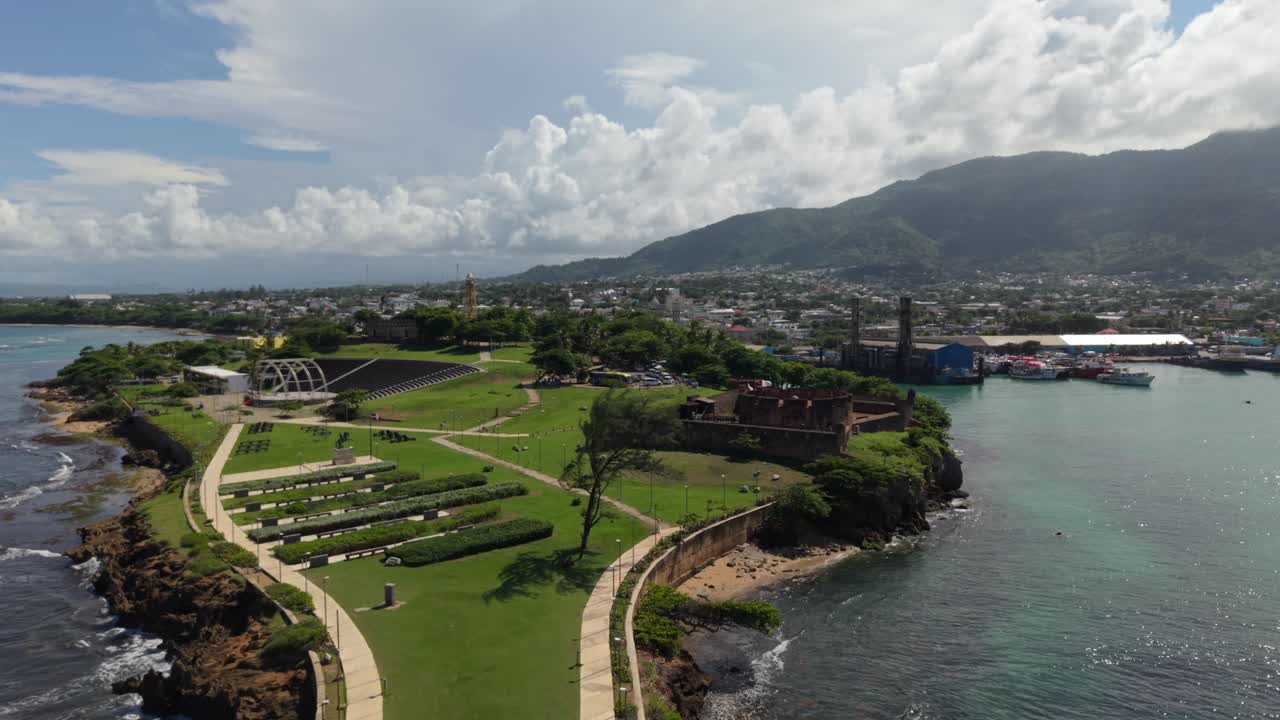 Ripresa aerea ad alta angolazione con allontanamento che rivela la Fortezza di San Felipe, punto di riferimento storico, e il paesaggio costiero urbano a Puerto Plata, con montagne e una baia sotto un cielo sereno.
