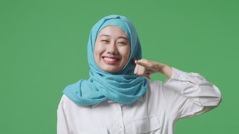primer plano de una mujer musulmana asiática sonriendo y haciendo un gesto de llamada mientras está de pie en el estudio de fondo de la pantalla verde
