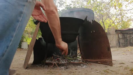 Man-Puts-Branches-In-The-Fire-While-Cooking-Fish-Stew-In-A-Pot-Outdoors