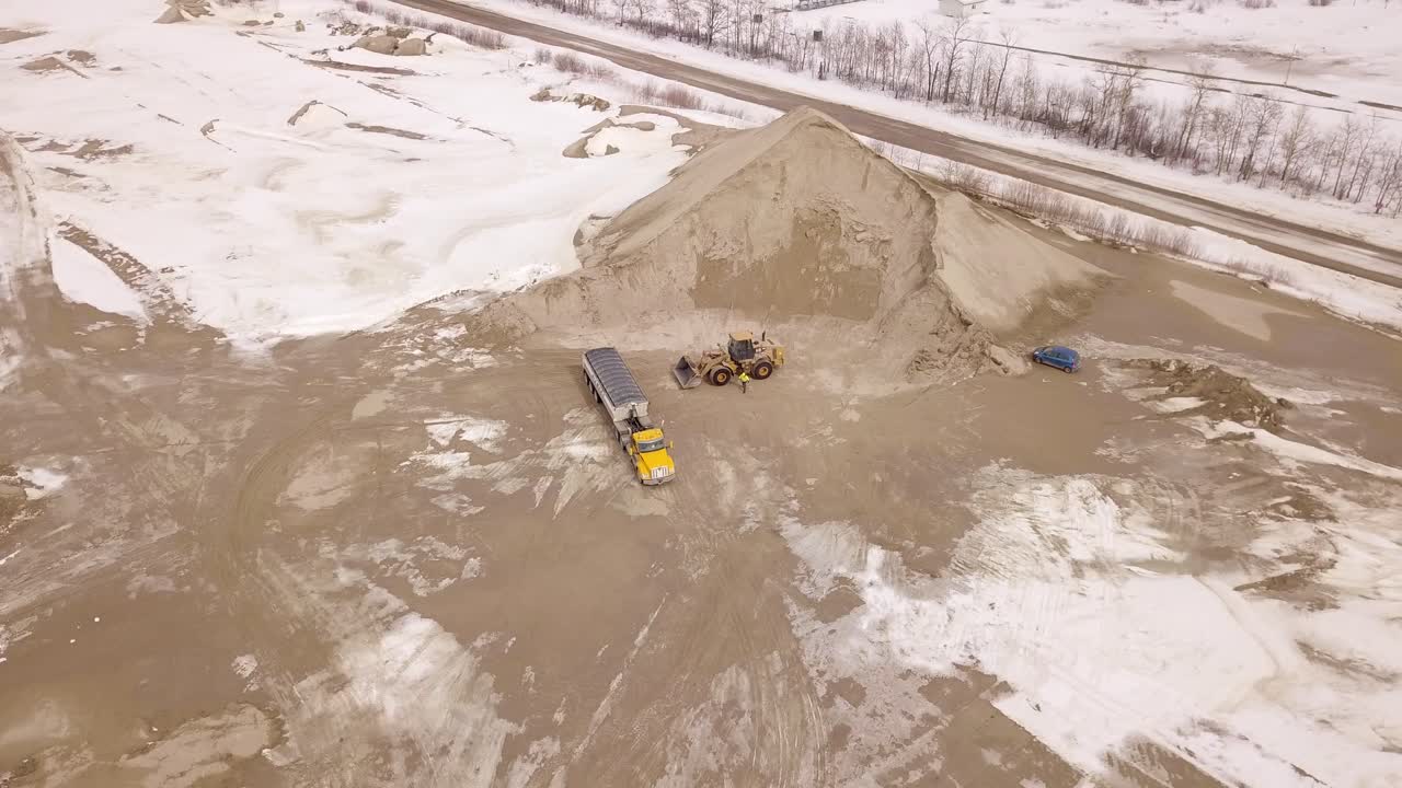 A wide drone shot showcases the full worksite as a truck and an excavator collaborate to transport and load sand efficiently.