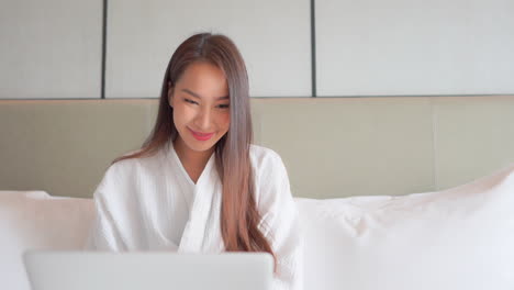Hübsche-Asiatische-Frau-Im-Weißen-Bademantel,-Die-Auf-Einem-Laptop-Computer-Tippt,-Der-Auf-Dem-Bett-Des-Hotelzimmers-Sitzt,-Gesicht,-Nahaufnahme,-Büste