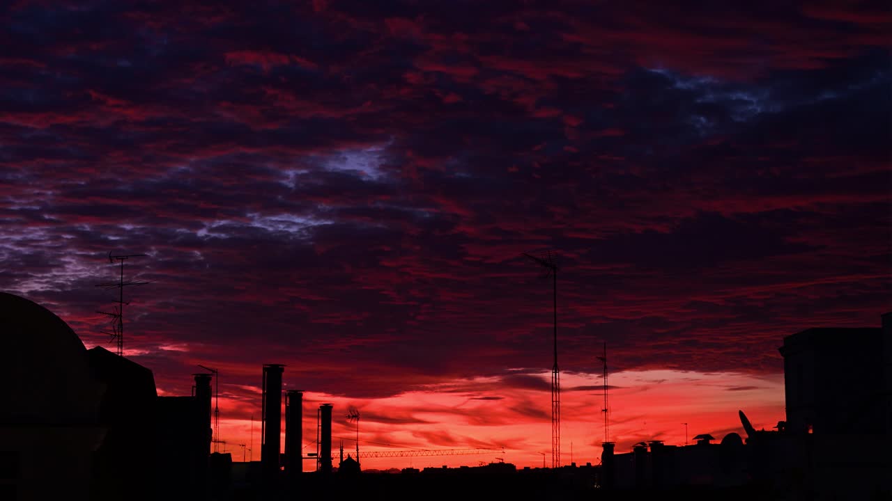 A wide shot of a colorful sunset sky over Madrid, Spain. The view includes the silhouettes of rooftops, chimneys, and antennas against a beautiful, cloudy sky at dusk