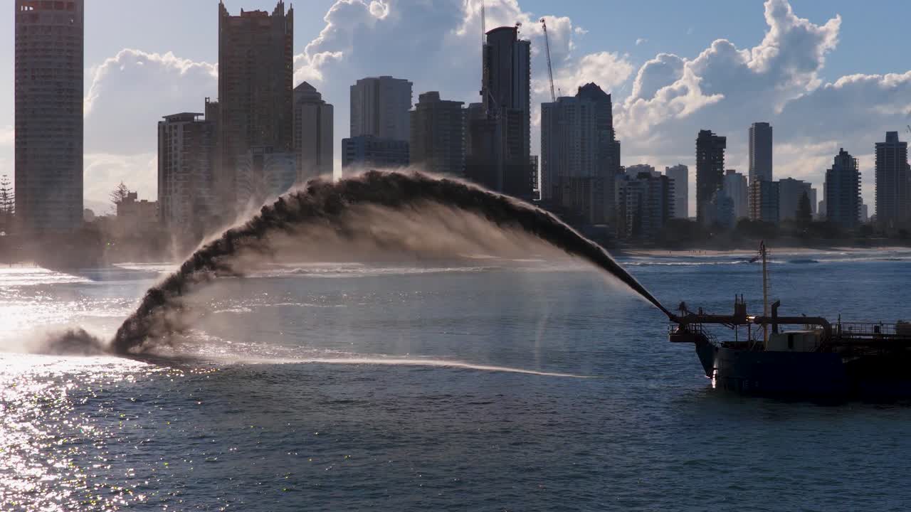 A dredging ship pumps sand onto a beach against a city skyline, under bright sunlight and clear skies