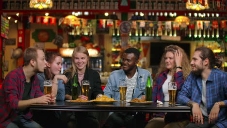 african american with friends at the bar drinks beer and eats chips friends raise glasses and bottles and knock / check on the table smiling and saying toasts.