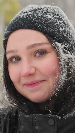 joven sonriendo cálidamente a la cámara en un parque nevado, sosteniendo una guitarra acústica roja vibrante, con la nieve descansando en su gorra negra y chaqueta, rodeada de árboles helados