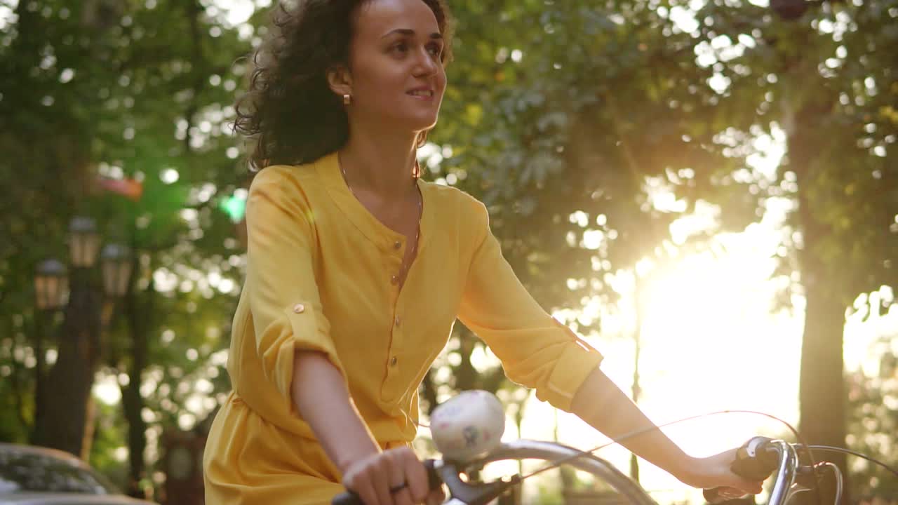 Lens flare: smiling happy woman in long yellow dress is riding a city bicycle with a basket and flowers in the park with green