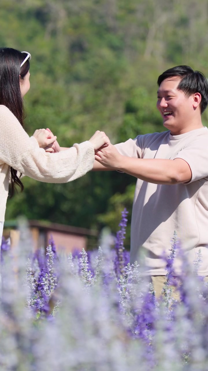 Pasangan tersenyum berpegangan tangan dengan ceria di ladang bunga ungu yang cerah, bidikan medium genggam.