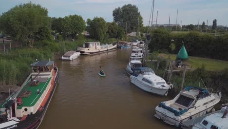 man kayaking on meandering river in-between boats, drone flies up as kayaker paddles towards camera