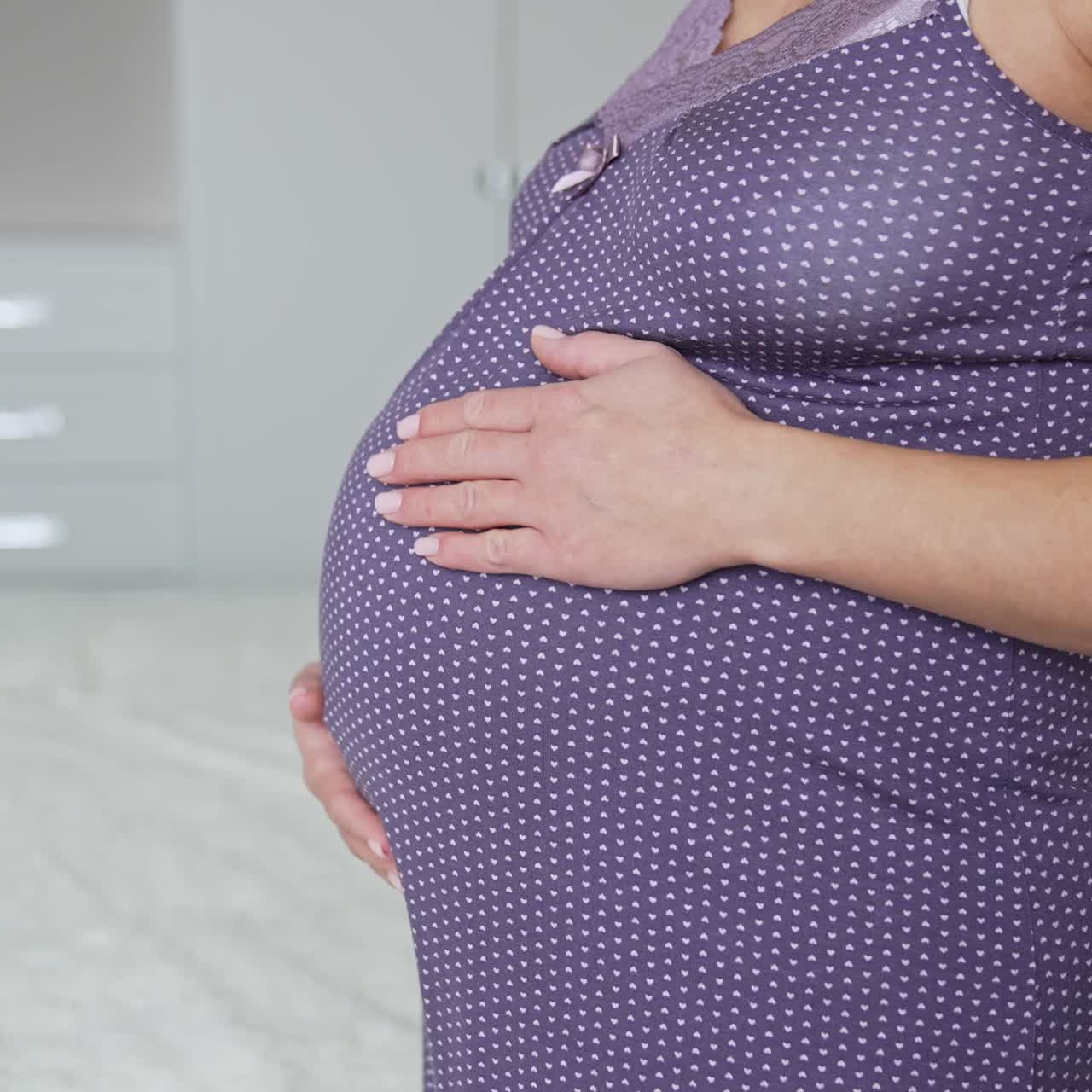 Mamma in attesa in camicia da notte in piedi in camera da letto. La donna accarezza teneramente la sua grande pancia con entrambe le mani. Primo piano.