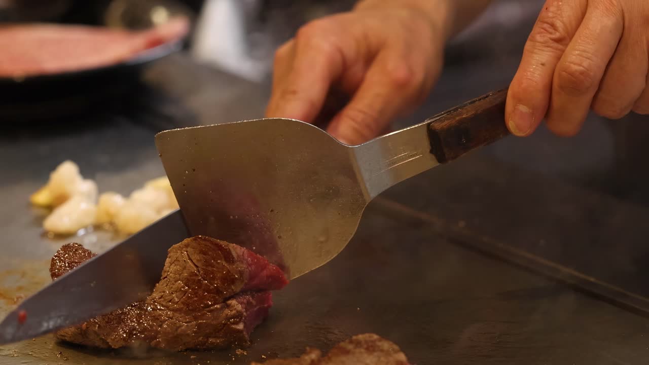Close-up of a chef using metal spatulas to flip and cook steak on a sizzling grill.
