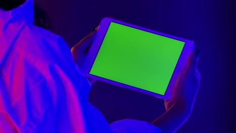 view from behind an african american woman holding a white tablet in her hand and touching a green screen chroma key. close up of a gadget against a background of bright neon lights. slow motion