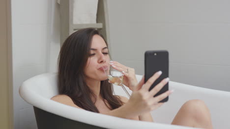 mujer acostada y relajada en el baño en casa posando para selfies en el teléfono móvil y bebiendo un vaso de vino