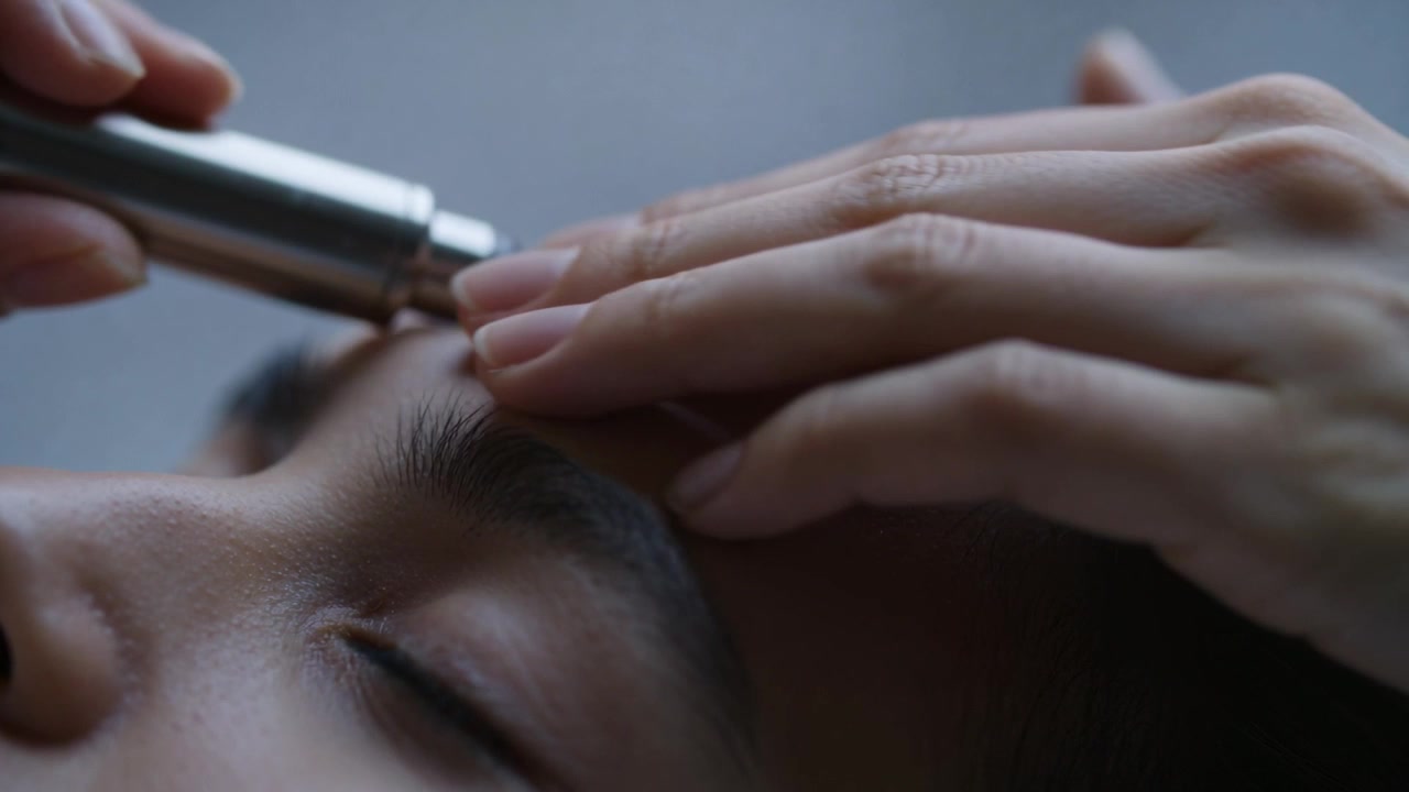 Approaching brow, device moving along right brow, hands pressing forehead at pillow for grooming